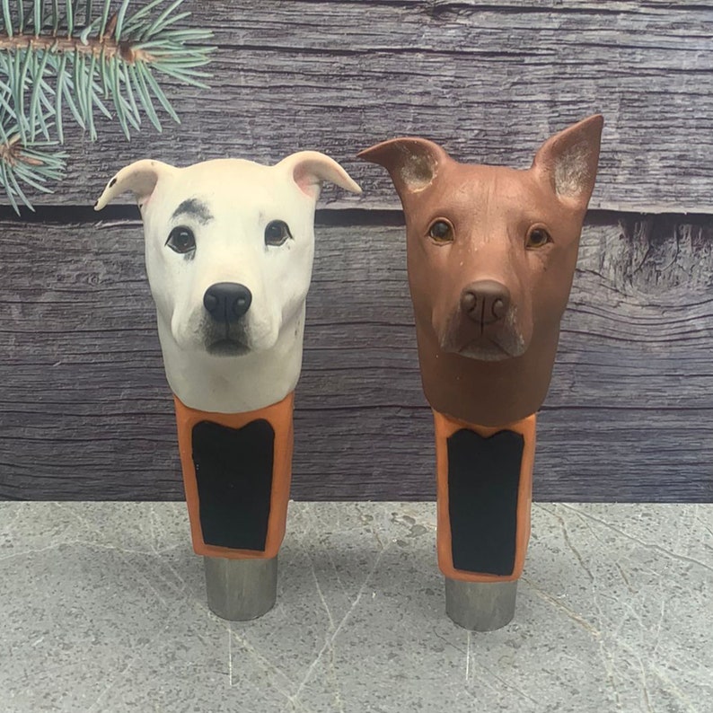 May include: Two ceramic dog head beer tap handles. The handle on the left is white with black spots and the handle on the right is brown. Both handles have a black chalkboard sign.