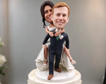 May include: A custom cake topper featuring a figurine of a person in a dark suit giving a piggyback ride to a person in a white wedding dress. The figures have large heads and are mounted on a circular white base atop a frosted cake.