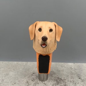Może przedstawiać: Ceramiczna klamka do kranu piwnego przedstawiająca realistyczny wizerunek głowy golden retrievera. Klamka jest pomalowana w odcieniach brązu i beżu, z czarnym prostokątnym miejscem na tekst lub logo.