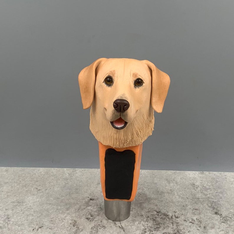 Może przedstawiać: Ceramiczna klamka do kranu piwnego przedstawiająca realistyczny wizerunek głowy golden retrievera. Klamka jest pomalowana w odcieniach brązu i beżu, z czarnym prostokątnym miejscem na tekst lub logo.