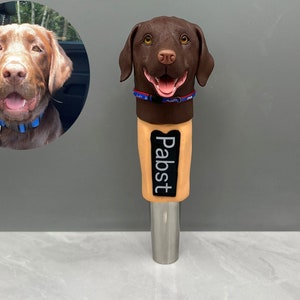 May include: A brown dog head beer tap handle with the word "Pabst" printed on the front. The tap handle is made of a brown material and has a silver metal base.