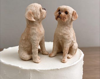 May include: Two light tan dog figurines with textured fur detailing are positioned on top of a round white frosted cake. One dog is facing the side while the other faces forward, both featuring dark eyes, noses, and small mouths.