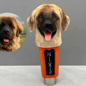 May include: A custom-made beer tap handle featuring a dog's head. The handle is made of brown and tan colored resin and has a black nose and tongue. The handle is attached to a metal base and has a black chalkboard with the name "Nixie" written on it.