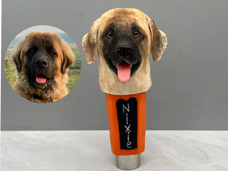 May include: A custom-made beer tap handle featuring a dog's head. The handle is made of brown and tan colored resin and has a black nose and tongue. The handle is attached to a metal base and has a black chalkboard with the name "Nixie" written on it.