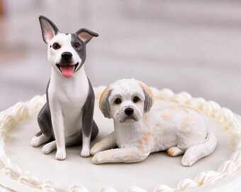 May include: Two dog figurines placed on top of a white frosted cake. One figurine is a grey and white dog sitting upright with its mouth open, while the other is a white and tan dog lying down in a resting position on the cake surface.