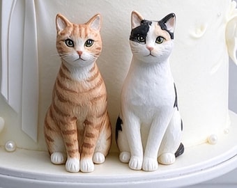 May include: Two cat figurines sitting upright on a white cake. One cat has orange tabby stripes with white paws, while the other is a white cat with black and orange patches. Both figurines have detailed facial features and are positioned side by side.