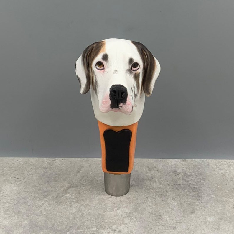 May include: A white, black, and brown ceramic beer tap handle with a dog's head. The dog has brown eyes and a black nose.