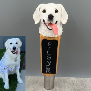 May include: A white ceramic dog head sculpture with a tongue sticking out, mounted on a wooden beer tap handle. The handle is labeled "PILSNER".