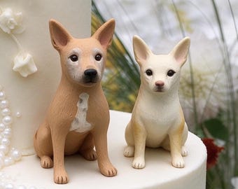 May include: Two dog figurines sitting on a white cake. The larger dog is tan with a white chest patch and dark nose. The smaller dog is cream-colored with pointed ears and dark eyes. Both figures are positioned upright on the smooth surface.
