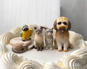 May include: A set of animal figurines on a white frosted cake, including a yellow and green parrot, a sleeping tan cat, a Siamese cat, a grey tabby cat, and a fluffy brown and tan dog, all arranged on the cake's surface.