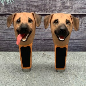 May include: Two brown and tan dog head shaped beer tap handles. The dog heads have black noses and black eyes. The tap handles are made of a ceramic material and have a black and orange design.