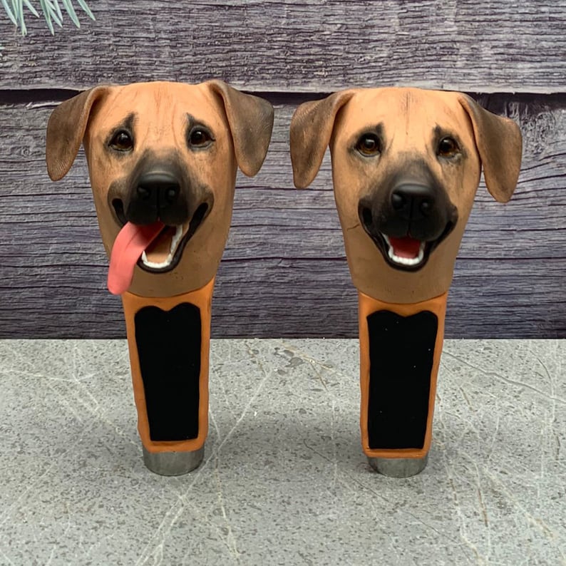 May include: Two brown and tan dog head shaped beer tap handles. The dog heads have black noses and black eyes. The tap handles are made of a ceramic material and have a black and orange design.