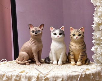 May include: Three cat figurines sitting on a white frosted cake. The left figure is a tan hairless cat, the center is a white cat with gray patches, and the right is a brown tabby cat with dark stripes. Each cat has detailed facial features.