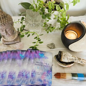 May include: A set of oracle cards with a purple and blue galaxy design. The cards are laid out on a white surface with a small white owl figurine, a candle holder, a sage bundle, and a multi-colored crystal wand. The card on top reads "Self Love & Inner Unity OR  Finding Divine Union as Within, So Without".