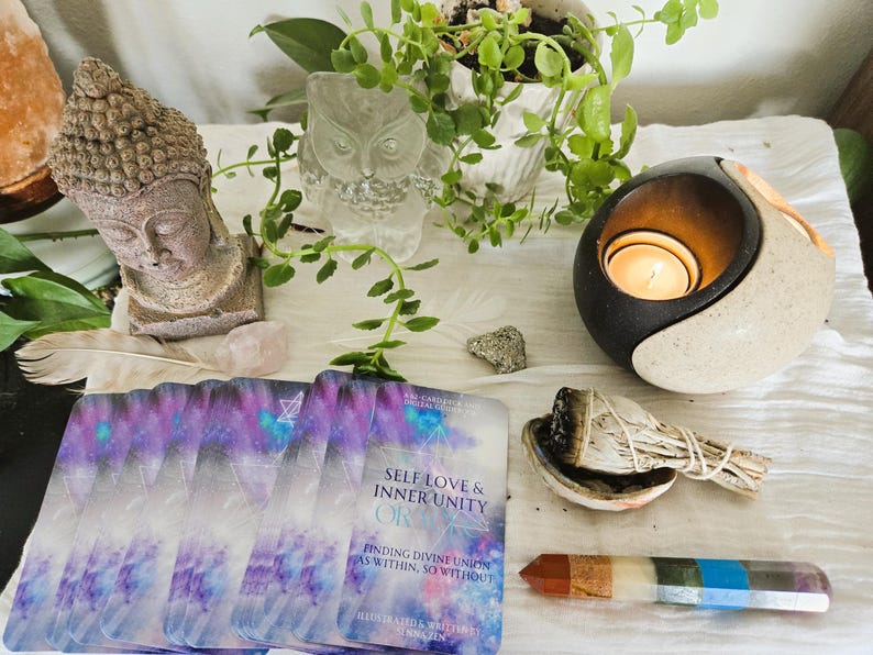 May include: A set of oracle cards with a purple and blue galaxy design. The cards are laid out on a white surface with a small white owl figurine, a candle holder, a sage bundle, and a multi-colored crystal wand. The card on top reads "Self Love & Inner Unity OR  Finding Divine Union as Within, So Without".