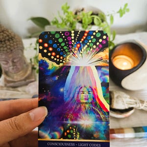 May include: A colorful card with a woman's face in the center. The background is a galaxy with a bright light shining from the center. The text on the card reads "Consciousness - Light Codes I Expand with an Open Heart and Mind".
