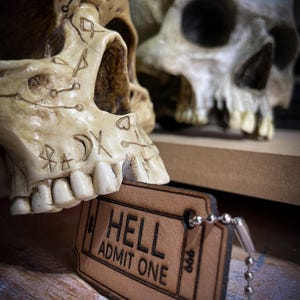 Może przedstawiać: Dwie białe ludzkie czaszki z misternymi zdobieniami, jedna z skórzanym breloczkiem w kształcie biletu z napisem "HELL ADMIT ONE".