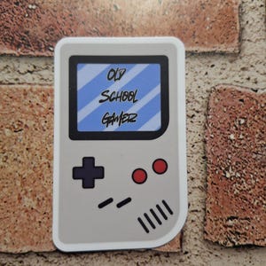 May include: A gray sticker in the shape of a retro handheld gaming device. The screen displays the text "OLD SCHOOL GAMER" in white. The device has a black directional pad, red buttons, and a speaker grill.