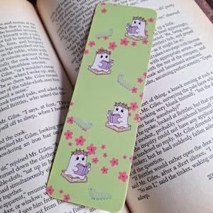 May include: A green bookmark featuring a repeating pattern of a cute ghost reading a book, wearing a flower crown, and a green caterpillar. Pink flowers are scattered across the bookmark.