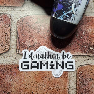 May include: A white sticker with the text "I'd rather be GAMING" in black, with a controller graphic. A video game controller with a black base and a white and blue design is partially visible. The background is a brick wall.
