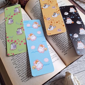 May include: Four colorful bookmarks with unique designs are displayed open on a book. The bookmarks feature illustrations of a ghost reading, a seashell on a float, and other whimsical designs. The bookmarks are rectangular and vary in color.