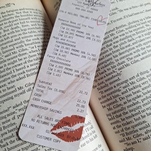Op de afbeelding: Een kassabon met tekst zoals "Romance Book of the Year" en "Hugs and Kisses." De bon heeft een lippenstiftkusafdruk en de datum 14-02-2023. Hij ligt op de open pagina's van een boek.