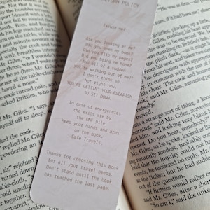 Op de afbeelding: Een crèmekleurig bladwijzer met tekst, rustend op de open pagina's van een boek. De bladwijzer heeft een 'Retourbeleid' met humoristische zinnen. De pagina's van het boek zijn verouderd, met zichtbare tekst.