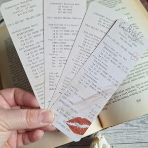 Op de afbeelding: Een hand houdt verschillende boekhandelbonnen vast, met tekst en prijzen. Eén bon heeft een lippenstiftkus en de tekst "ALL SALES ARE FINAL". De bonnen liggen op een boek.