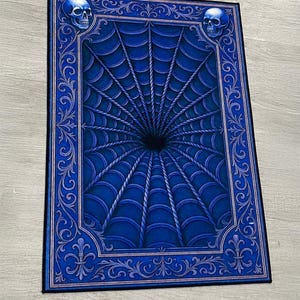 May include: A rectangular rug featuring a deep blue color palette with a central optical illusion of a dark tunnel surrounded by a web-like pattern. The border is decorated with ornate scrollwork and two skulls at the top corners. Text reads Sample.