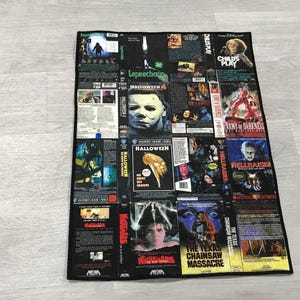 May include: A rectangular fabric throw blanket featuring a collage of classic horror movie VHS cover art. The print includes titles such as Halloween, Child's Play, Hellraiser, Nightmare on Elm Street, and The Texas Chainsaw Massacre with black borders.