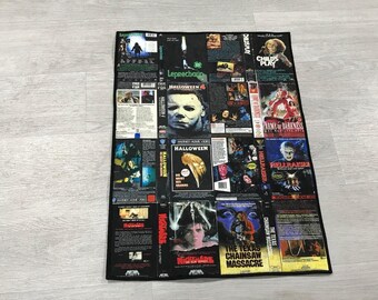 May include: A rectangular fabric throw blanket featuring a collage of classic horror movie VHS cover art. The print includes titles such as Halloween, Child's Play, Hellraiser, Nightmare on Elm Street, and The Texas Chainsaw Massacre with black borders.