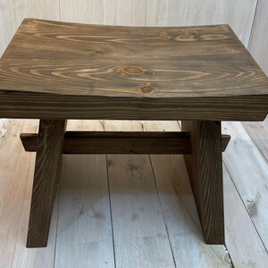 K&ouml;nnte beinhalten: Ein dunkelbrauner Holzhocker mit einer abgerundeten Oberseite und einem einfachen, robusten Design. Der Hocker ist aus massivem Holz gefertigt und hat eine nat&uuml;rliche Oberfl&auml;che.