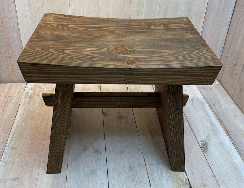 K&ouml;nnte beinhalten: Ein dunkelbrauner Holzhocker mit einer abgerundeten Oberseite und einem einfachen, robusten Design. Der Hocker ist aus massivem Holz gefertigt und hat eine nat&uuml;rliche Oberfl&auml;che.