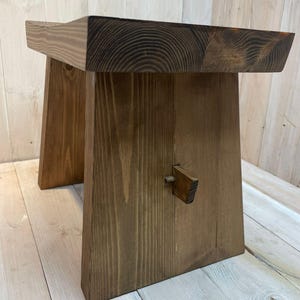 K&ouml;nnte beinhalten: Eine dunkelbraune Holzbank mit schlichtem Design. Die Bank hat eine rechteckige Oberseite und zwei schr&auml;ge Beine. Die Beine sind durch ein kleines Holzst&uuml;ck miteinander verbunden.