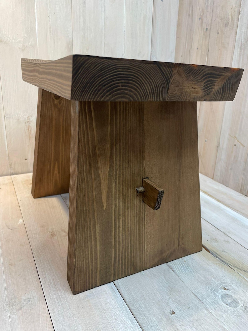 K&ouml;nnte beinhalten: Eine dunkelbraune Holzbank mit schlichtem Design. Die Bank hat eine rechteckige Oberseite und zwei schr&auml;ge Beine. Die Beine sind durch ein kleines Holzst&uuml;ck miteinander verbunden.