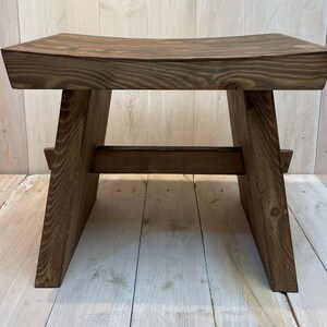 K&ouml;nnte beinhalten: Ein dunkelbrauner Holzhocker mit einer gew&ouml;lbten Sitzfl&auml;che und einem einfachen, robusten Design. Der Hocker hat eine rechteckige Basis mit zwei schr&auml;gen St&uuml;tzen auf jeder Seite.