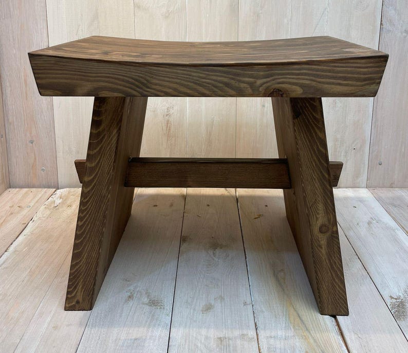 K&ouml;nnte beinhalten: Ein dunkelbrauner Holzhocker mit einer gew&ouml;lbten Sitzfl&auml;che und einem einfachen, robusten Design. Der Hocker hat eine rechteckige Basis mit zwei schr&auml;gen St&uuml;tzen auf jeder Seite.