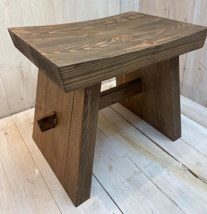 K&ouml;nnte beinhalten: Ein dunkelbrauner Holzhocker mit einer gew&ouml;lbten Oberseite und einem einfachen, robusten Design. Der Hocker hat ein einzigartiges, abgewinkeltes Bein-Design und ein kleines Holzst&uuml;ck am vorderen Bein.