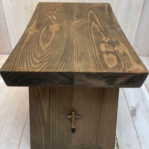 K&ouml;nnte beinhalten: Ein dunkelbrauner Holzhocker mit einem einfachen, rechteckigen Design. Die Oberseite des Hockers besteht aus einem einzigen St&uuml;ck Holz mit einer nat&uuml;rlichen Maserung. Der Hocker hat ein kleines, dekoratives Metallkreuz auf der Vorderseite.
