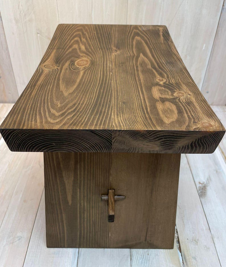 K&ouml;nnte beinhalten: Ein dunkelbrauner Holzhocker mit einem einfachen, rechteckigen Design. Die Oberseite des Hockers besteht aus einem einzigen St&uuml;ck Holz mit einer nat&uuml;rlichen Maserung. Der Hocker hat ein kleines, dekoratives Metallkreuz auf der Vorderseite.