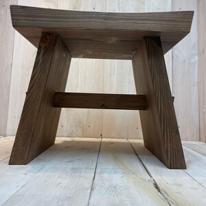 K&ouml;nnte beinhalten: Ein dunkler brauner Holzhocker mit einem einfachen, rechteckigen Design. Der Hocker hat eine breite, flache Oberseite und einen stabilen Sockel mit abgewinkelten Beinen.