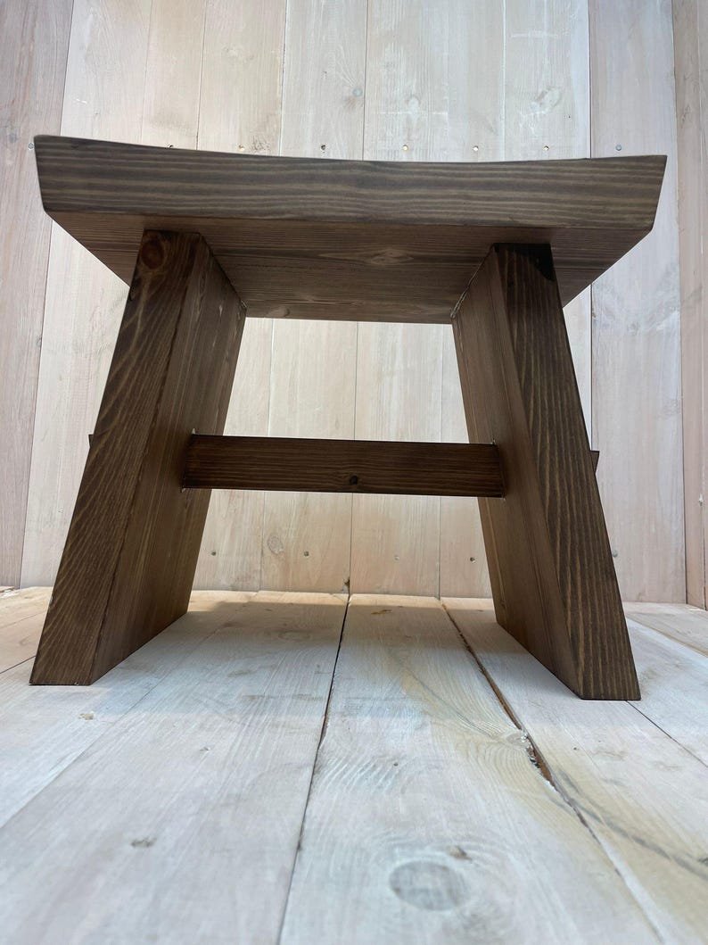 K&ouml;nnte beinhalten: Ein dunkler brauner Holzhocker mit einem einfachen, rechteckigen Design. Der Hocker hat eine breite, flache Oberseite und einen stabilen Sockel mit abgewinkelten Beinen.