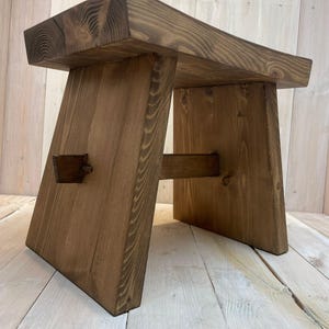 K&ouml;nnte beinhalten: Ein Holzhocker mit einer dunkelbraunen Beize. Der Hocker hat eine rechteckige Oberseite und ein einzigartiges, abgewinkeltes Bein-Design.