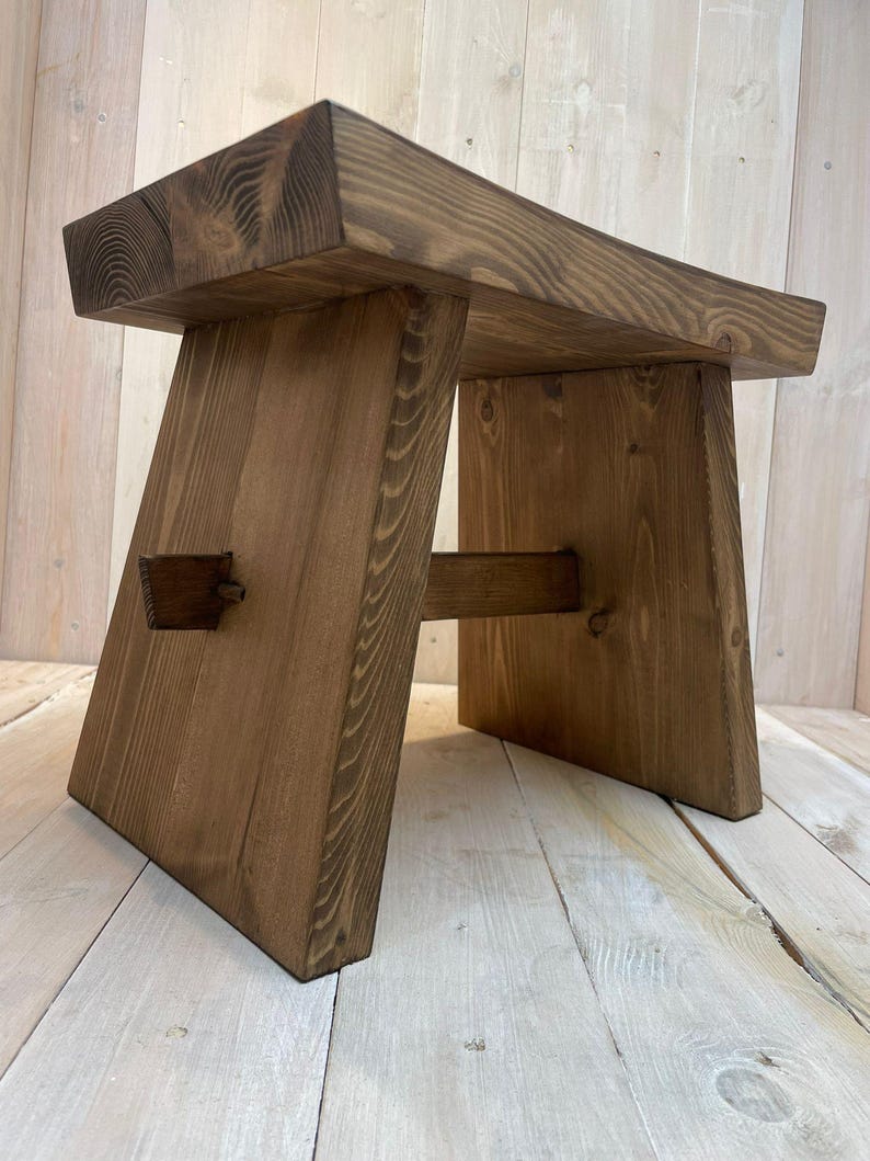 K&ouml;nnte beinhalten: Ein Holzhocker mit einer dunkelbraunen Beize. Der Hocker hat eine rechteckige Oberseite und ein einzigartiges, abgewinkeltes Bein-Design.