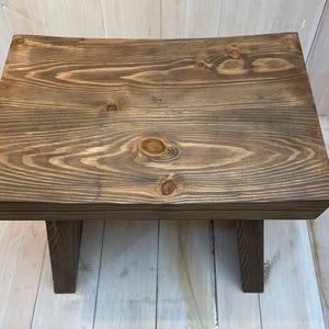 K&ouml;nnte beinhalten: Ein dunkelbrauner Holzhocker mit einer gew&ouml;lbten Oberseite und zwei kurzen Beinen. Der Hocker ist aus massivem Holz gefertigt und hat eine nat&uuml;rliche Holzmaserung.