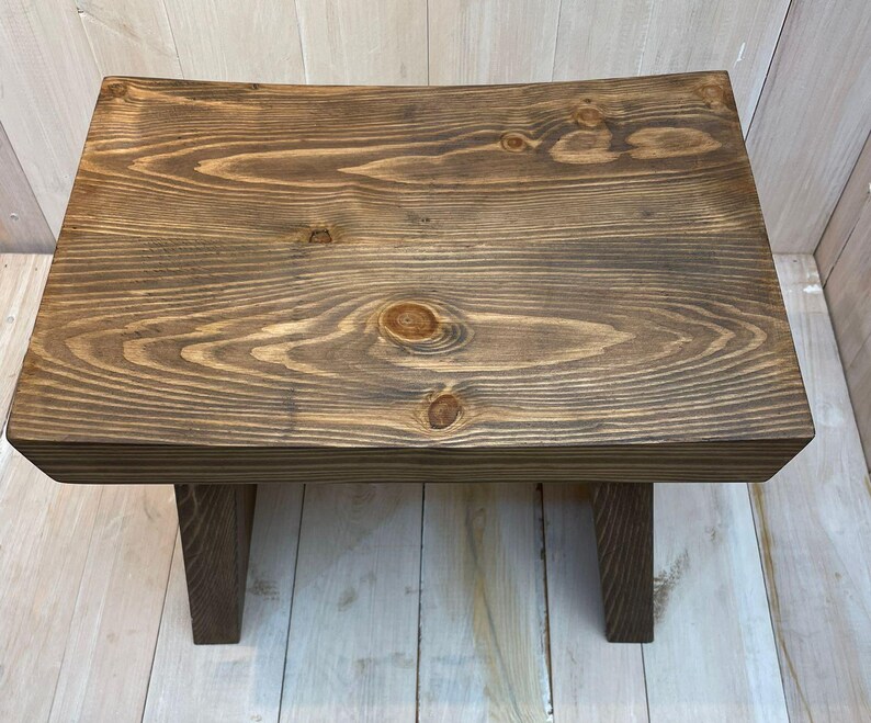 K&ouml;nnte beinhalten: Ein dunkelbrauner Holzhocker mit einer gew&ouml;lbten Oberseite und zwei kurzen Beinen. Der Hocker ist aus massivem Holz gefertigt und hat eine nat&uuml;rliche Holzmaserung.