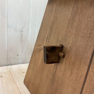 K&ouml;nnte beinhalten: Ein Holztritt mit brauner Beize. Der Hocker hat einen kleinen Holzkeil, der als Verriegelungsmechanismus dient.