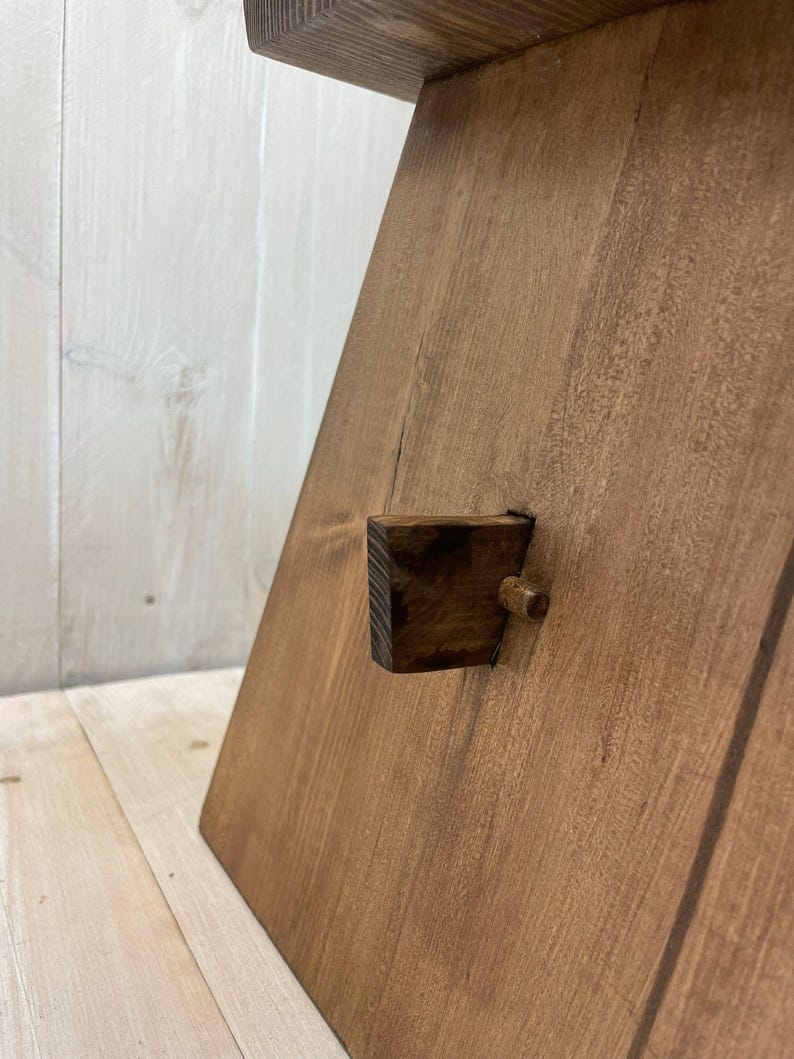 K&ouml;nnte beinhalten: Ein Holztritt mit brauner Beize. Der Hocker hat einen kleinen Holzkeil, der als Verriegelungsmechanismus dient.
