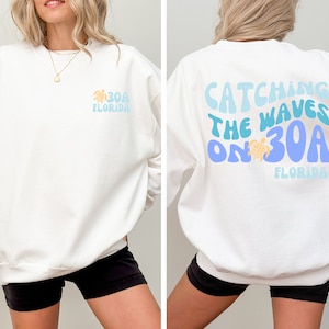 以下が含まれることがあります： 前面に「30A FLORIDA」、背面に「CATCHING THE WAVES ON 30A FLORIDA」と書かれた白いスウェットシャツ。デザインには、青と黄色の色合いのカメのグラフィックが含まれています。