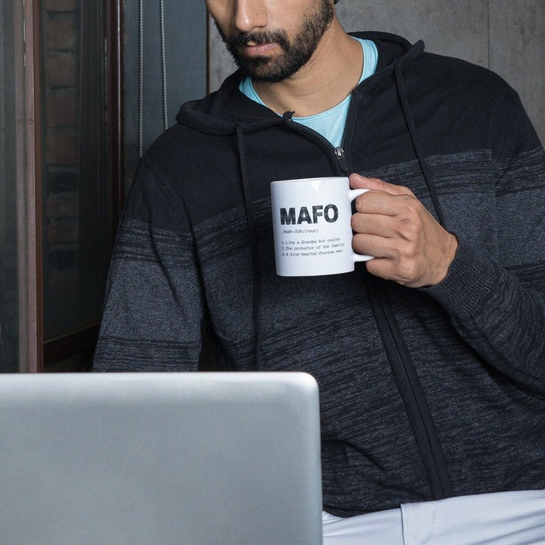 May include: White ceramic mug with the word "MAFO" printed in bold black letters. Below the word is a definition with three bullet points. The mug is held by a person wearing a black and gray striped zip-up hoodie.
